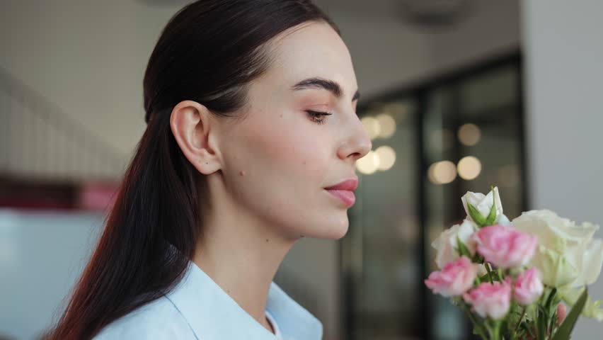 Happy young woman in a blue shirt is shown in profile, smelling a bouquet of pink and white roses and smiling. She is standing in a bright office space. Perfect for lifestyle, beauty.