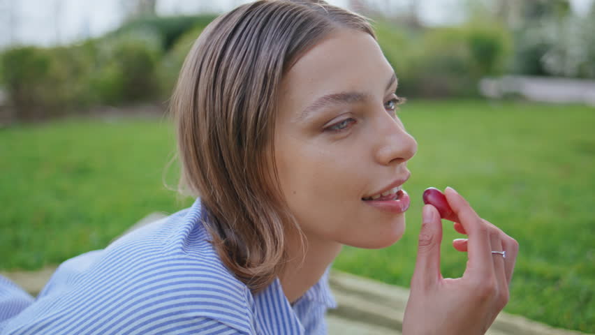 Eating girl enjoying picnic in green park closeup. Portrait of satisfied woman chewing grape on beautiful nature. Carefree brunette smiling tasting fresh healthy snack lying on blanket at vivid grass