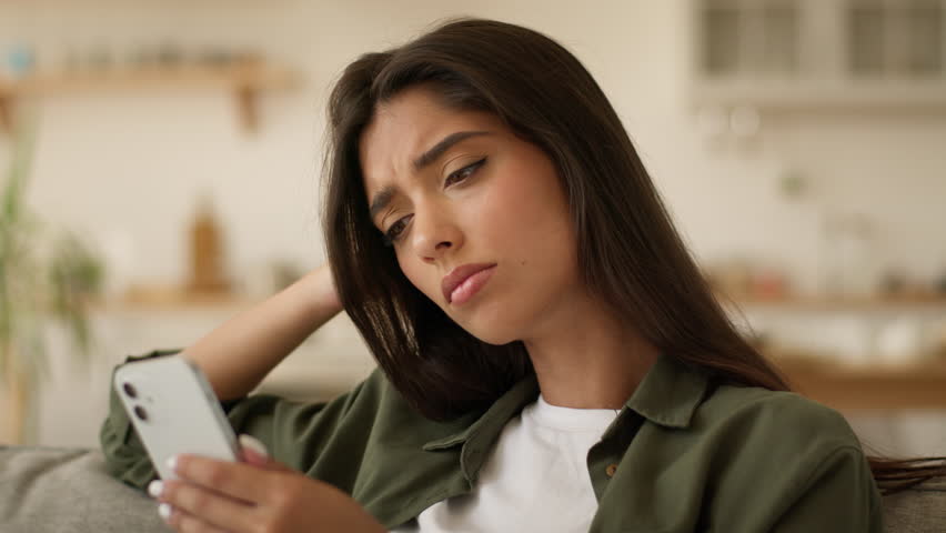 Upset sad frustrated disappointed confused puzzled Indian woman Arabian girl female Muslim lady at home couch sofa looking mobile phone smartphone social media app worry bad news failure lost online