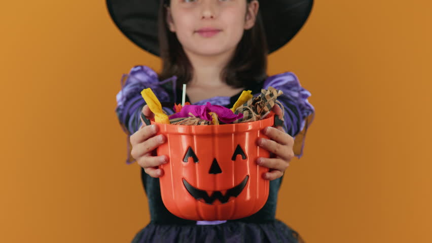Medium close-up of hands of young girl in black and purple sorceress or witch costume posing on orange backdrop, holding out pumpkin bucket full of Halloween candy after successful trick or treating