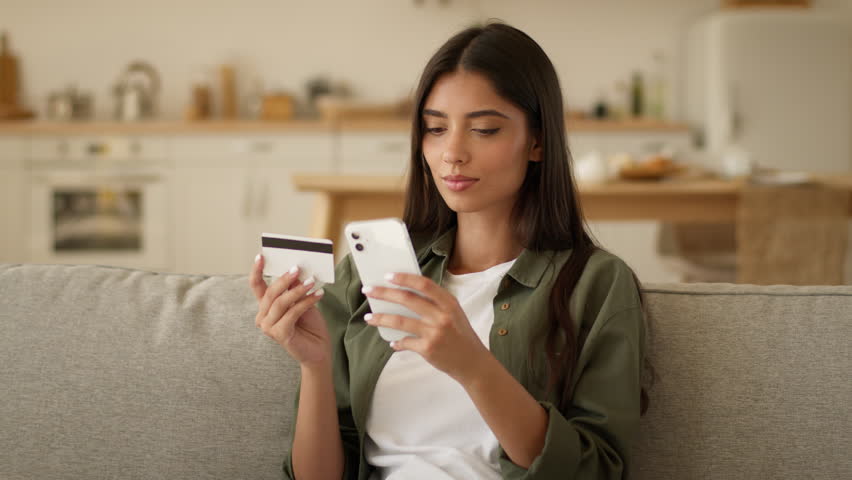 Happy smiling Indian woman Arabian girl client customer buyer female Muslim lady at home couch sofa kitchen shopping online mobile phone order food delivery enter number credit bank card buy purchase