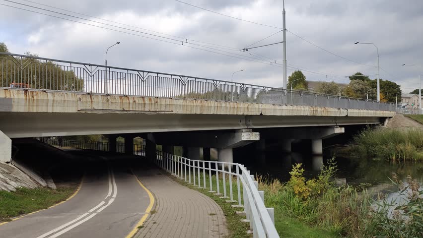 Above the river is a concrete bridge with metal fencing and light poles. Beyond the bridge are trees and buildings, and below it is a paved bicycle lane and tiled sidewalk. Cloudy autumn weather