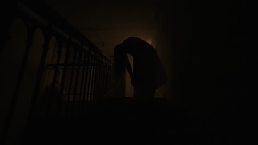 Creepy ghost woman in white dress and scary makeup standing alone with bended body on staircase in dark haunted house. Halloween horror advertisement concept.