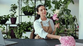 A female florist wearing a work apron sits in her flower shop surrounded by plants and flowers. She holds a colorful bouquet and looks at the camera smiling. - Powered by Shutterstock - Get 15% off with code: PIKWIZARD15