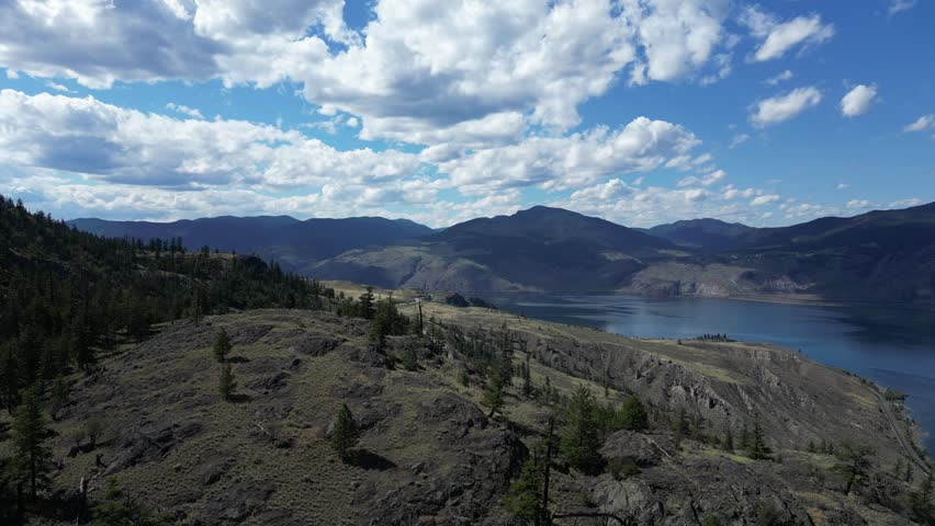 Winding mountain road carves through a rugged landscape, leading towards a serene lake nestled amidst the peaks. Blue sky and fluffy clouds, complete the picturesque scene. British Columbia, Canada.