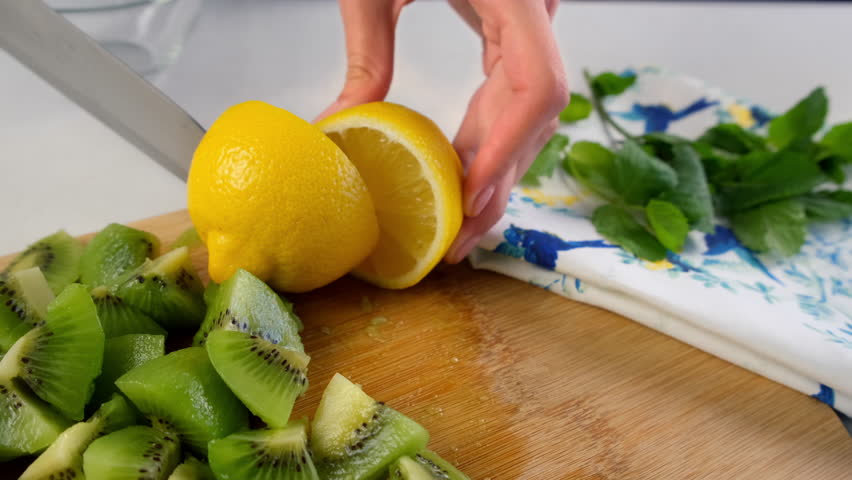 Woman cutting lemon on halves on wooden board with kiwi and mint leaves cooking fruit lemonade on kitchen at home. Preparing ingredient for dish or beverage. Recipe cuisine professional culinary.