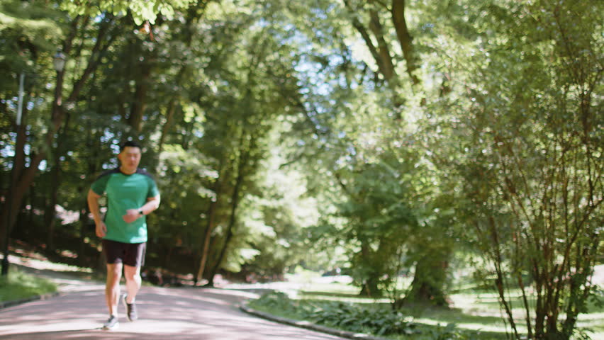 Young adult Asian runner having heart attack or chest pain after running workout in park on sunny summer day. Tired Japanese athlete suffers from tension in the heart. Health care and sports concept.