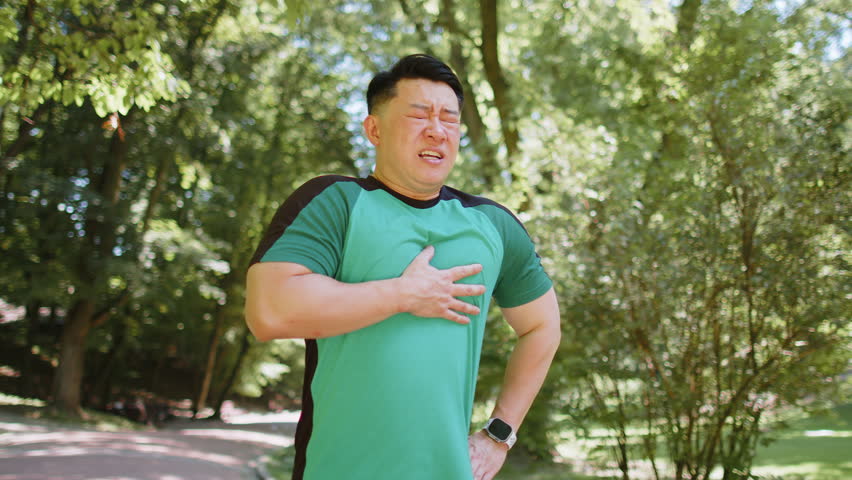 Young adult Asian runner having heart attack or chest pain after running workout in park on sunny summer day. Tired Japanese athlete suffers from tension in the heart. Health care and sports concept.