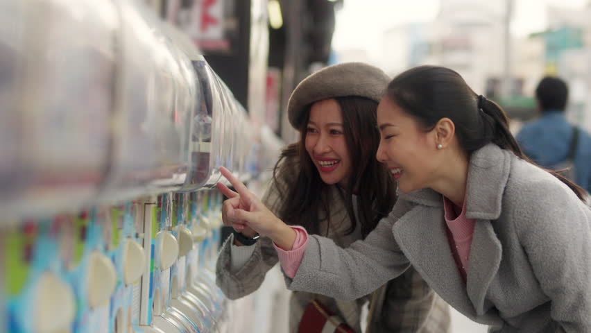 Asian woman friends handle a doll from gashapon capsule toy vending machine while shopping together at Tokyo city, Japan. Attractive girl enjoy and fun outdoor travel city street on autumn vacation.