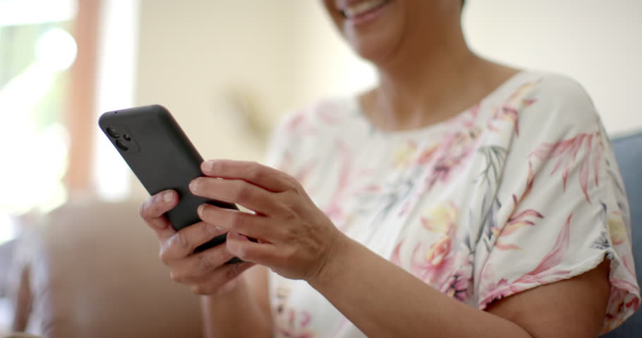 Happy senior biracial woman sitting on couch and talking on smartphone at home, slow motion. Domestic life, technology, retirement and lifestyle, unaltered.