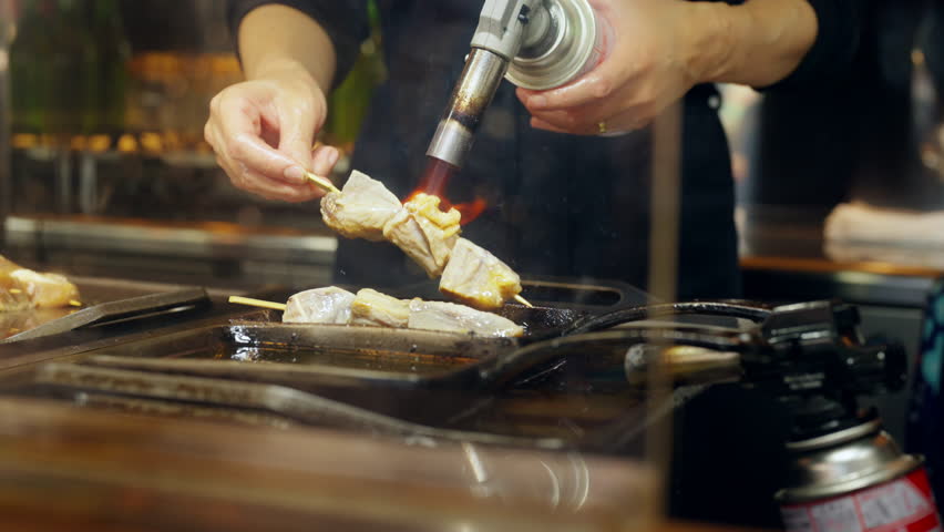 4K Chef hand using torch gas burner cooking grilled seafood tuna meat bbq on sticks serving to customer at street market in Tokyo city, Japan. Japanese street food and holiday travel vacation concept.
