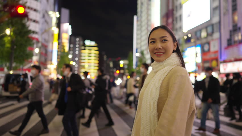 Happy Asian woman travel walking, shopping in the city on holiday vacation. Attractive girl enjoy urban outdoor lifestyle crossing city street crosswalk with crowd of people in Tokyo, Japan at night.