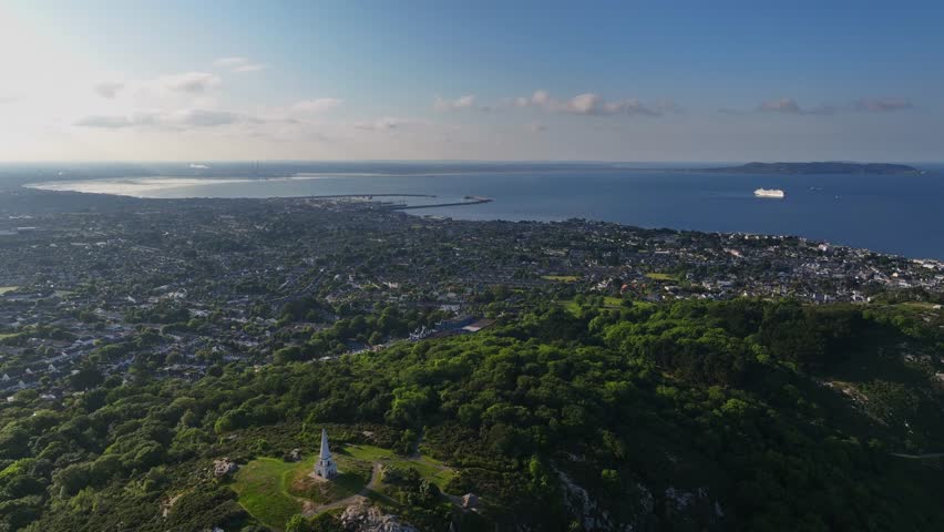 Killiney Hill, County Dublin, Ireland, June 2023. Drone gradually descends towards The Obelisk and pushes north west with Dún Laoghaire harbour and the city center in the distance on a sunny day.