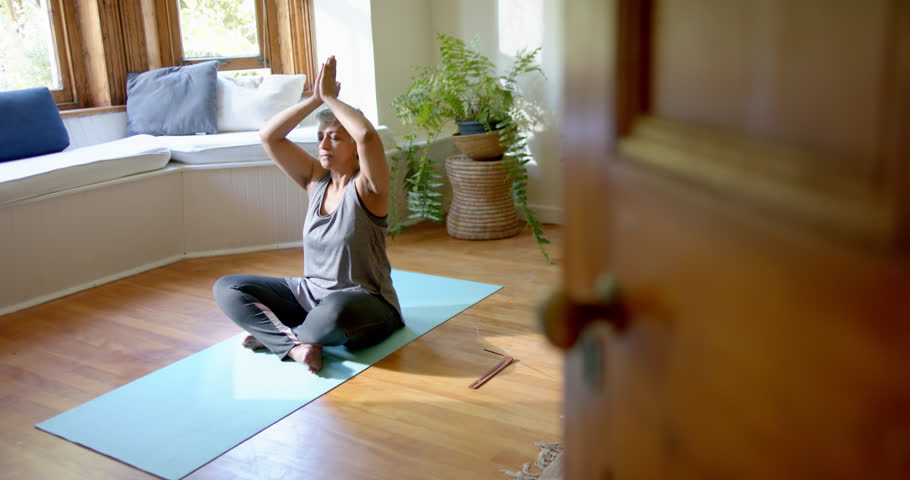 Focused senior biracial woman meditating on yoga mat at home. Domestic life, yoga, lifestyle, unaltered.