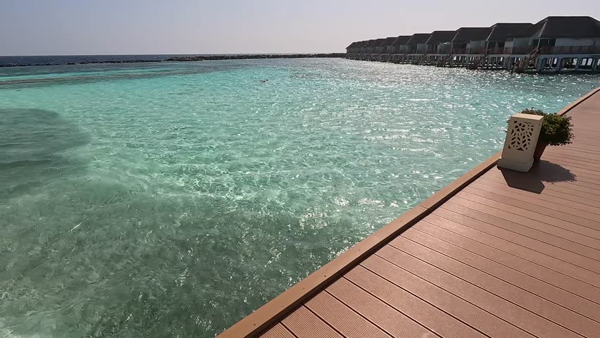 View of the Maldives island lagoon from a wooden bridge. Crystal-clear water with a school of fish, sandy beach.