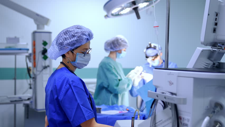 Female anesthesiologist stands looking at the apparatus in surgery room. Surgeon and scrub nurse work at operation at backdrop. - Powered by Shutterstock - Get 15% off with code: PIKWIZARD15