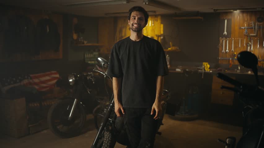 Portrait of a happy brunette mechanic guy with stubble in a gray T-shirt who poses against the background of tools and motorcycles in his workshop studio