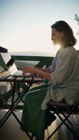 A young Caucasian woman is working on a laptop while sitting on a balcony with a beautiful view during sunset.