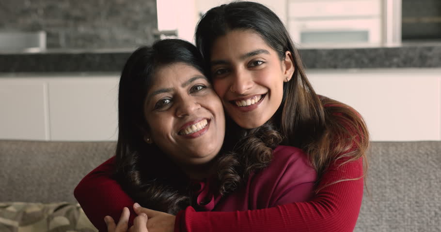 Happy cheerful adult Indian daughter girl embracing cheerful senior mom from behind, looking at camera with toothy smile. Young woman hugging middle aged mother with love, affection, support