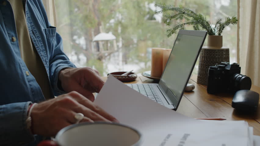 Side tilt shot of male Caucasian writer working on laptop, looking through printed version of new autobiography book and drinking hot tea, sitting by window in cottage house during winter holidays