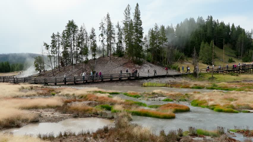 A slow-motion clip of a group of numerous tourists walking on a boardwalk that passes by a natural hot pool in the geyser basin area in the Yellowstone valley.