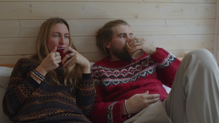 Medium shot of calm middle-aged woman and bearded man in sweaters drinking hot tea and looking out window. Spouses resting together in country house on winter holidays