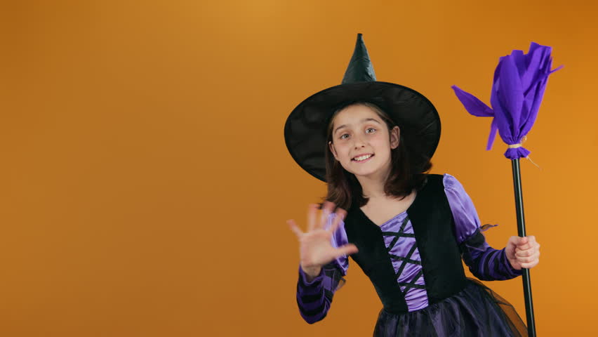 Medium full looped footage of happy young Caucasian girl in purple witch dress, black hat, holding broom, appearing in corner on orange backdrop, smiling, waving at camera, disappearing. Copy space