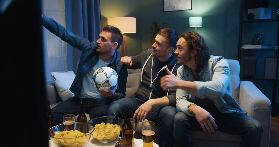 Three handsome Caucasian guys best friends sitting on couch in evening with football ball, snacks and beer and taking selfie photo with smartphone camera. Sport fans posing to phone at home at TV.