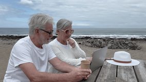 Happy senior couple sitting at table on beach using laptop surfing together, shopping, video chatting. Elderly enjoying authentic retirement lifestyle using modern technology. Horizon over sea - Powered by Shutterstock - Get 15% off with code: PIKWIZARD15