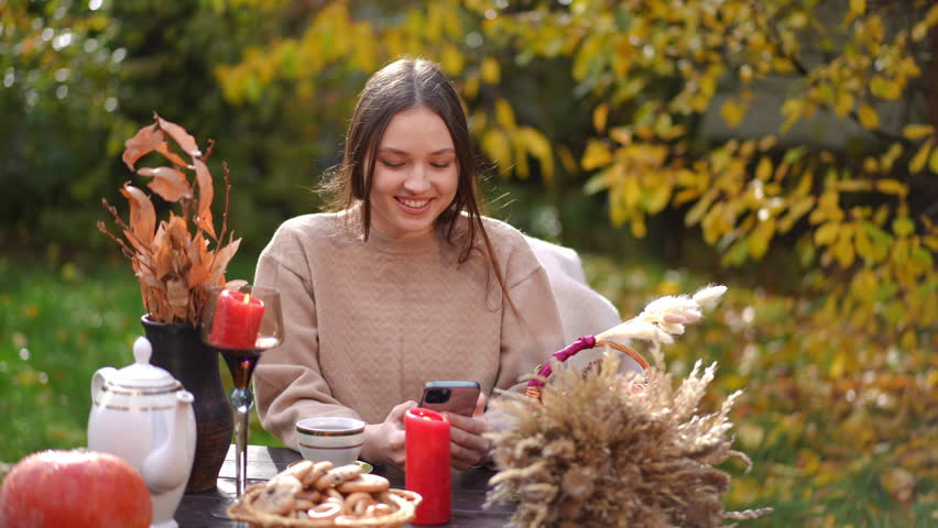 A cheerful woman smiles at an autumn gathering, enjoying her smartphone and festive snacks, creating joyfulness