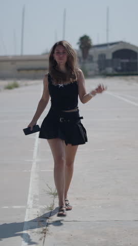 young girl caucasian woman in short black dress walking facing camera happy and content while listening to music and dancing with headphones on her ears