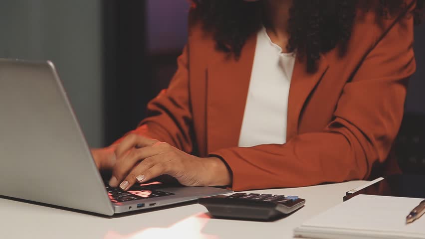 Careful businesswoman analyzing financial graphs and reports on her computer, working overtime to type out company strategies for an investment plan in a startup office