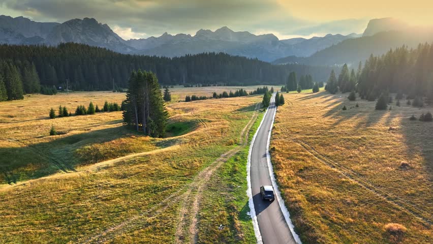 Aerial drone shot of car driving on scenic road in mountains. Flying over scenic road in the mountains with car moving along it. Sunset view of mountains road. Summer adventure by car