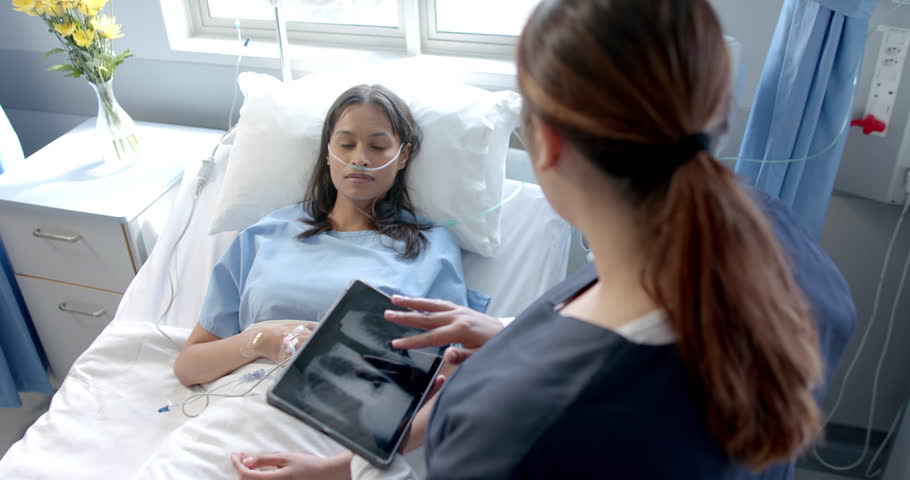Diverse female patient in hospital bed and female doctor using tablet talking, slow motion. Communication, consultation, treatment, care, hospital, work, medicine and healthcare, unaltered.