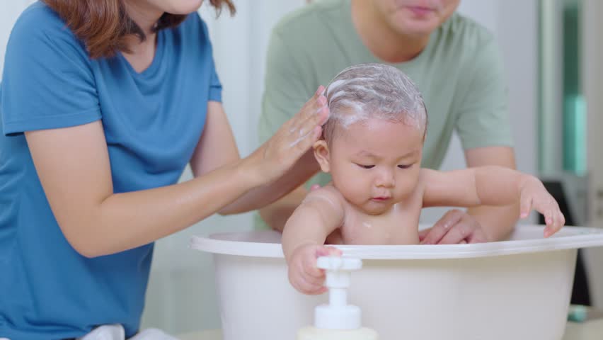 The father and mother are helping each other bathe their baby in a bathtub. In the picture, everyone looks happy.	