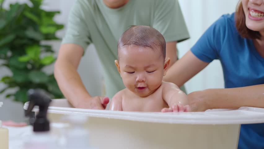 The father and mother are helping each other bathe their baby in a bathtub. In the picture, everyone looks happy.	