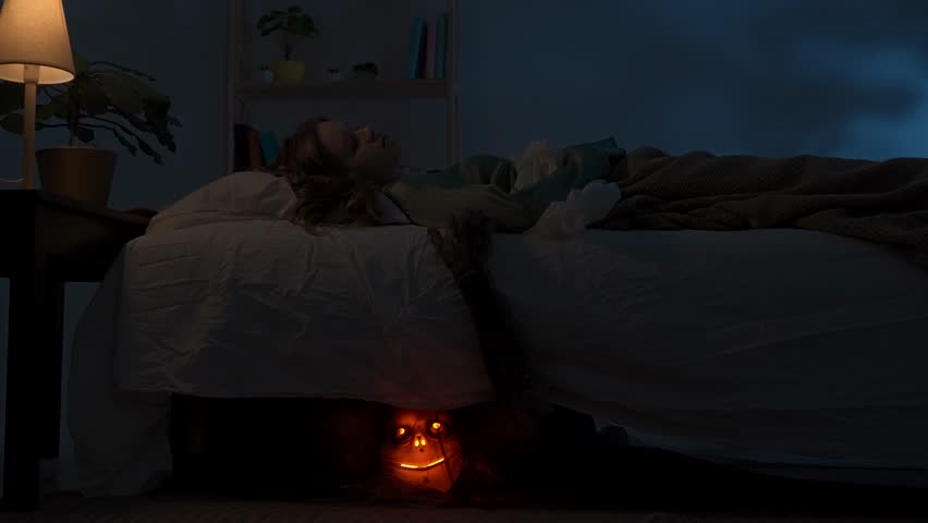 Woman sleeping in bed in dark apartment room, monster with scary face hiding under bed, creepy hand crawling out and touching girl. Halloween horror concept.