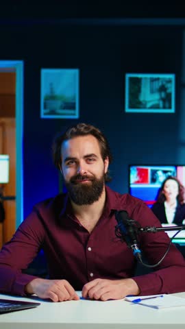 Vertical video Joyful content creator sitting at table talking in microphone in dimly lit home studio interior, chatting with subscribers. Upbeat man surrounded by podcast recording gear in apartment