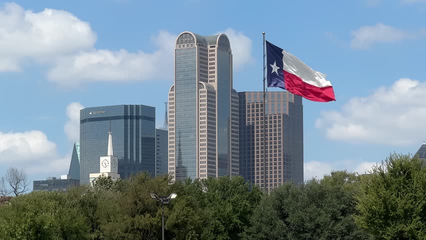 Dallas, TX USA - September 21, 2024: Handheld wide-shot video of Texas flag blowing in the wind in front of the downtown Dallas skyline on the last hot day of summer 2024. Heatwaves can be seen in the