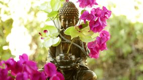 Seamless looping video: Water flows through a brass Buddha fountain decorated with an offering of pink flowers. Sunlight-dappled foliage is blurred in the background.