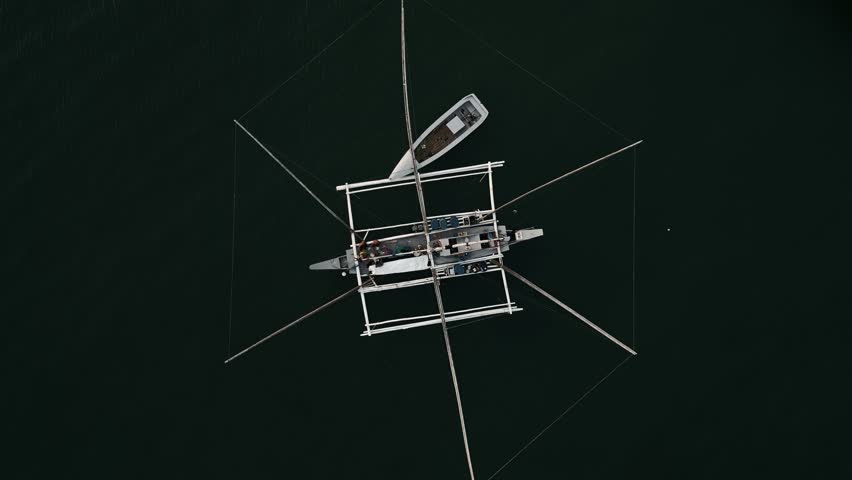 Top-down aerial orbit of double hull outrigger canoe with intricate network of fishing nets at Taloot Argao Port's pier and boats at sunrise in Cebu, Philippines, with calm waters