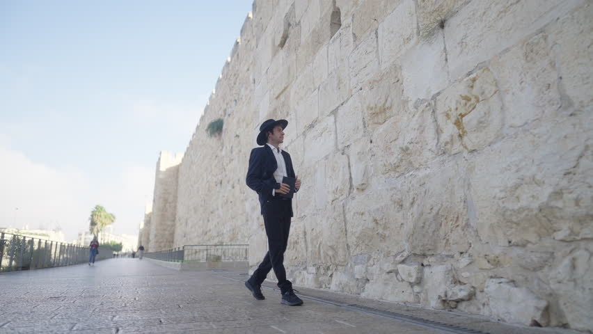 An Orthodox man in a black suit and hat walks down an alley by the walls of the Old City, also known as the City of David, in Jerusalem