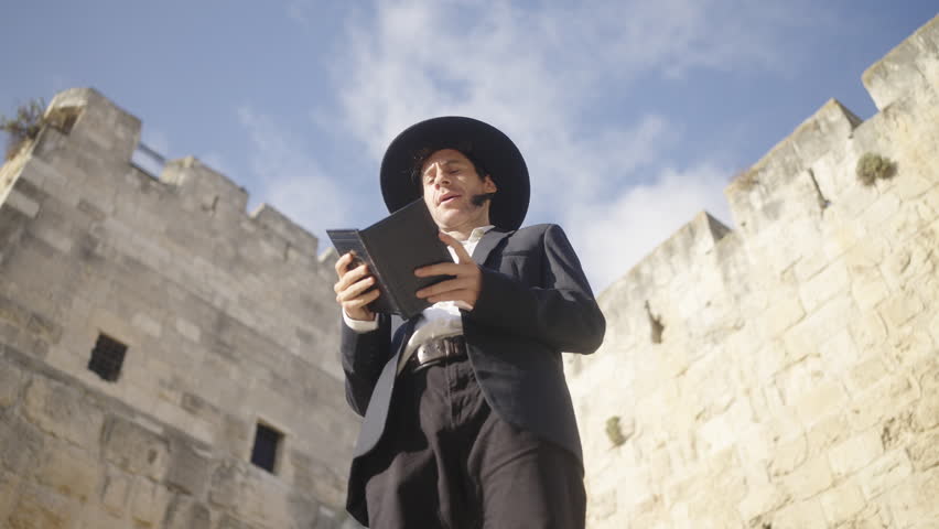 Pan shot of an Orthodox male in a black suit and hat, praying and reading from a Tehilim book beside an ancient old city in Jerusalem