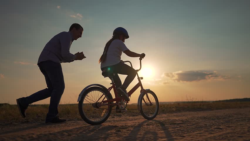 Father helps child ride bike at sunset. Child learning to balance while father supports. Playful family moment outdoors. Bike ride during sunset playtime. Father and child enjoy sunset bike play.