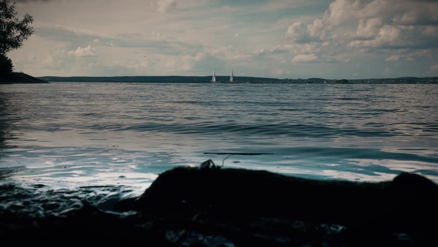 View from the darkened shore of two sailboats sailing on a blue lake. 