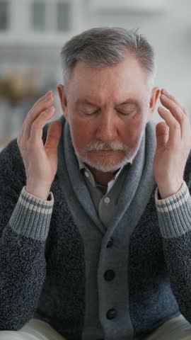 Senior man feeling headache, massaging temples, sitting on sofa in living room, vertical video. Migraine and hypertension in old age, relax and keep calm, single retiree male person resting at home
