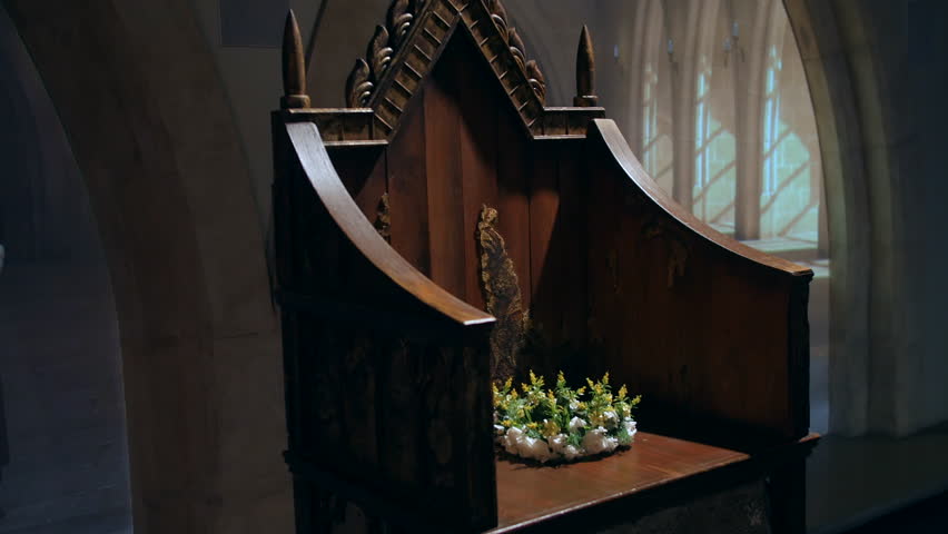 A historic wooden throne with detailed carvings. A floral wreath rests on the seat, emphasizing the throne's significance and the solemn atmosphere of memorial