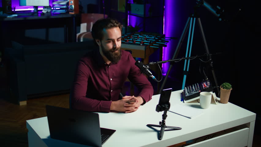 Media star talking in front on smartphone attached to tripod stabilizer in studio, discussing with viewers. Man using mobile phone to record video in living room for social media, camera B