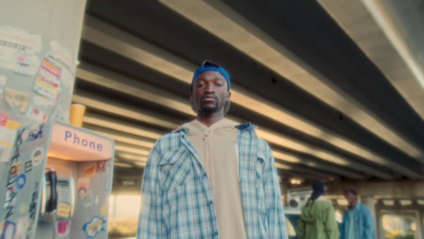 Young cool African American man wearing headphones over neck standing beside payphone under urban bridge, looking into camera with serious expression. Video portrait, whip zoom shot