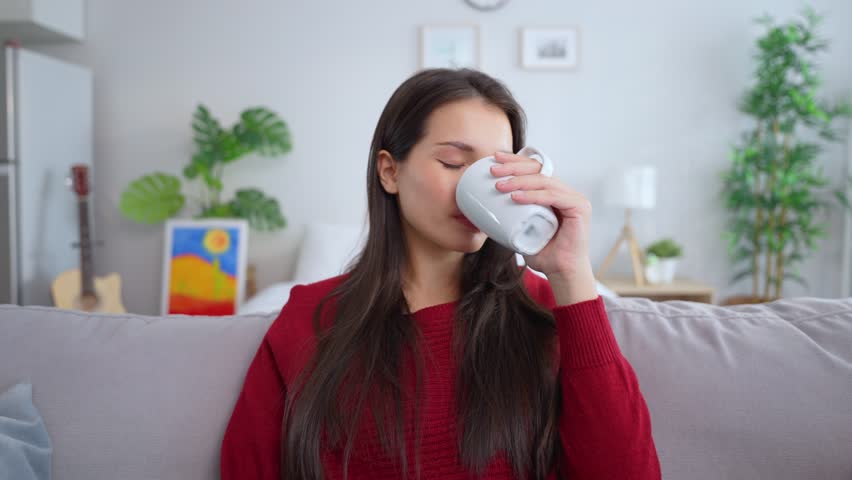 Portrait of Asian young woman drinking a cup of hot coffee in house. Attractive casual girl feel happy and relax, sitting on sofa and looking at camera while drink a mug of milk in living room at home